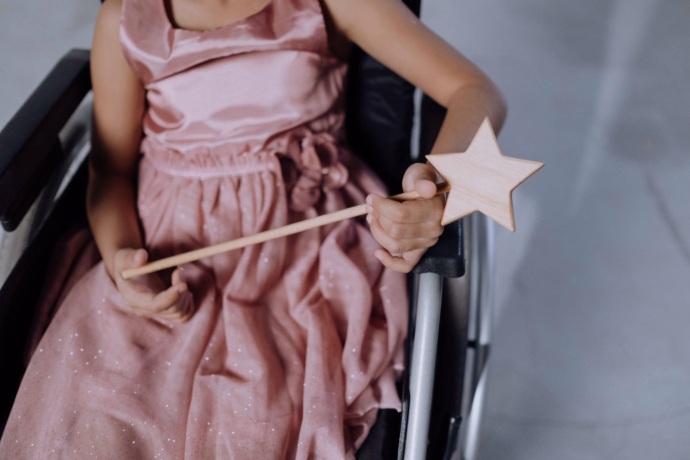 A child in a pink party dress seated in a wheelchair, holding a wooden star wand across her lap