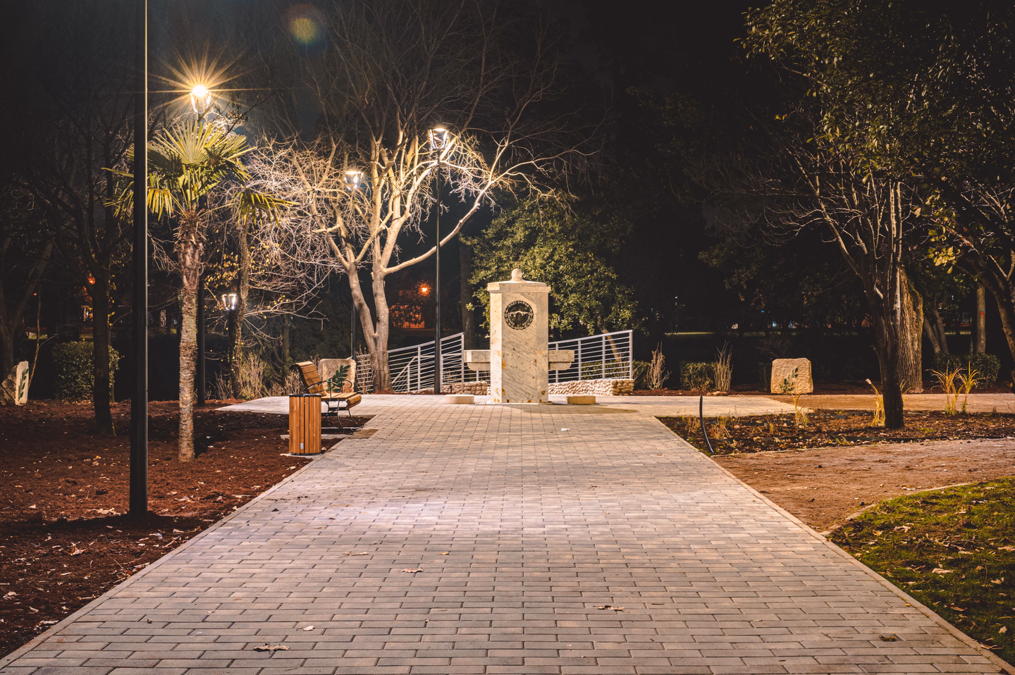 A wide paved path at night lit evenly by lamps, with bare trees on either side