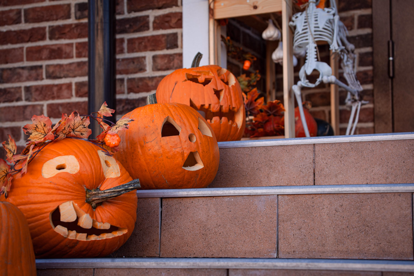Three carved jack-o'-lanterns sitting on concrete porch steps, with a skeleton standing by the door behind them