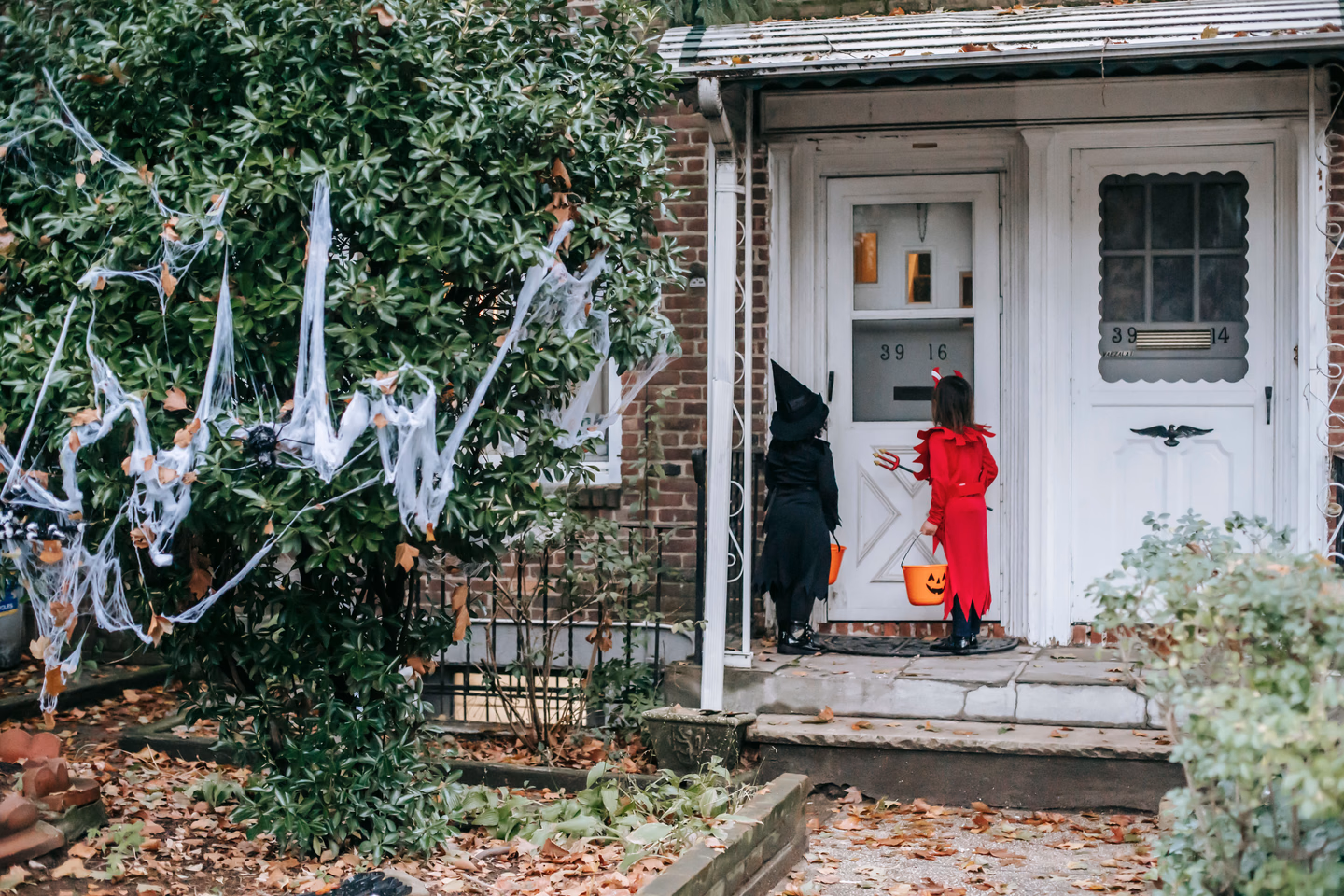 Two children in a black witch costume and a red devil costume, one holding a pumpkin bucket, waiting at a white front door with fake cobwebs strung over the bushes beside the steps