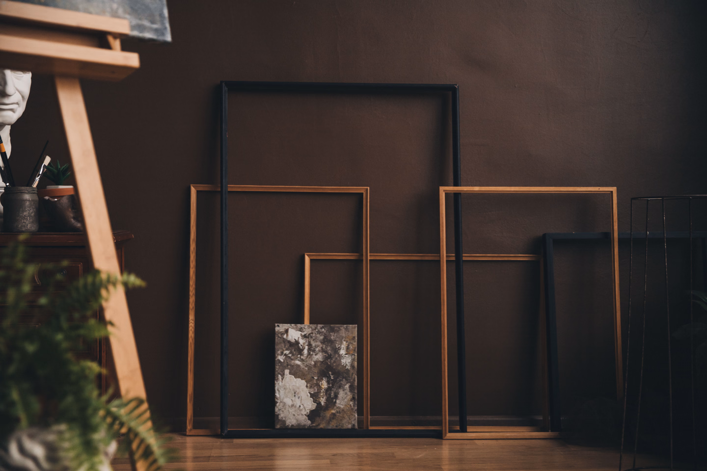 Several empty wooden picture frames of different sizes leaning against a dark brown wall in a dim studio