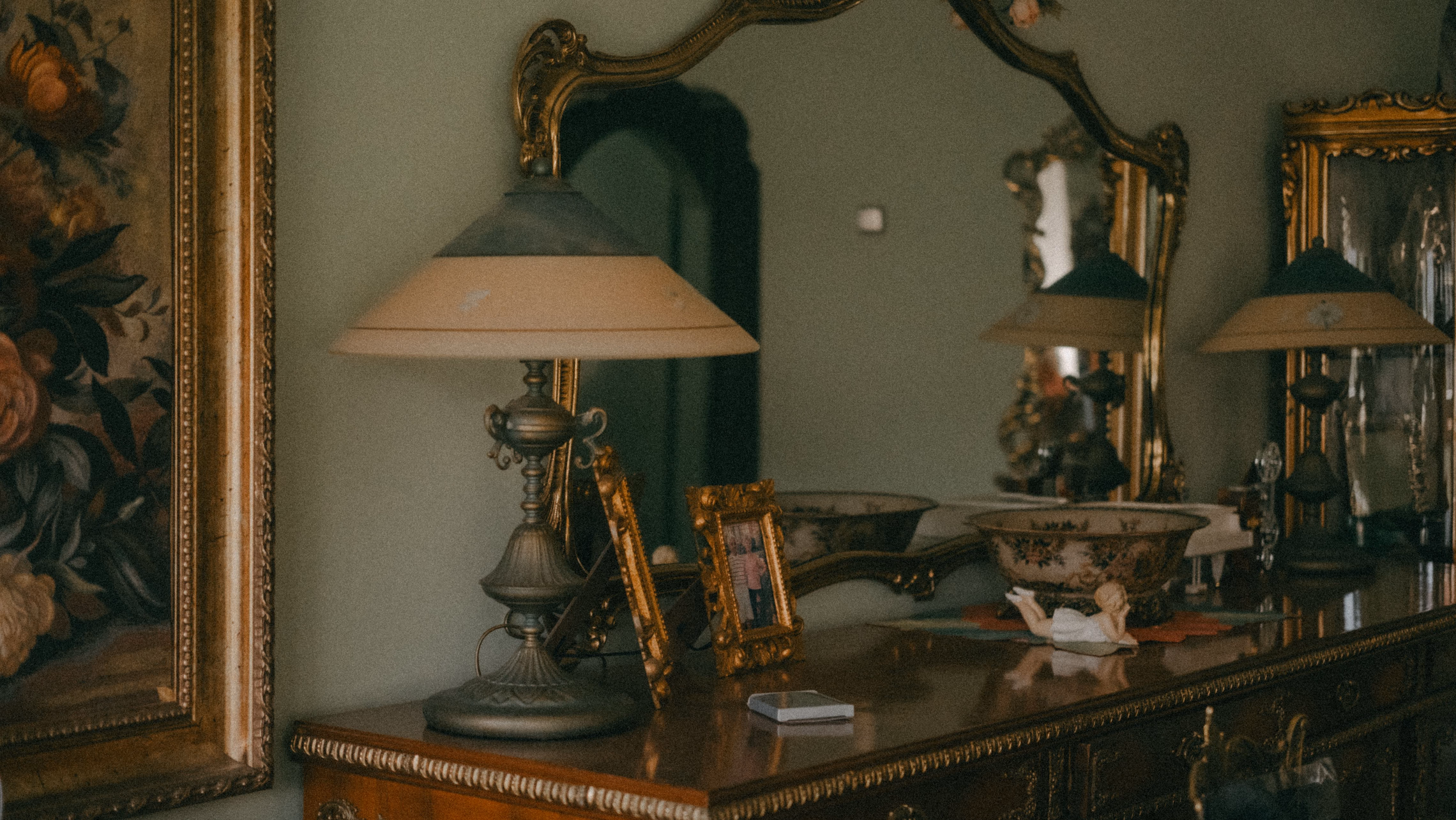 An old dresser with brass table lamps and small gilt photo frames in front of an ornate gold-framed mirror, in dim green light