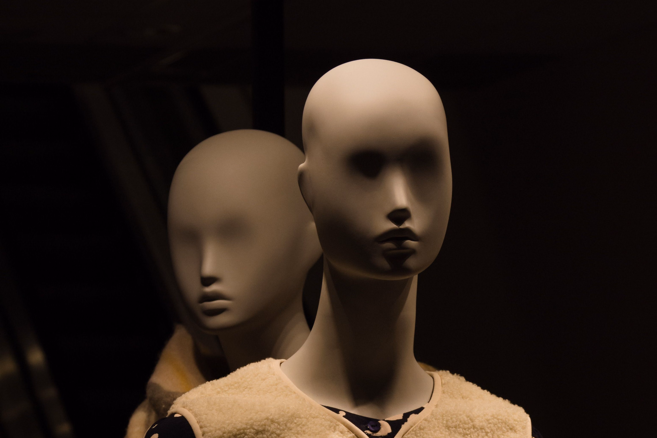 Two bald, featureless display mannequins in dim amber light, the nearer one wearing a shearling vest