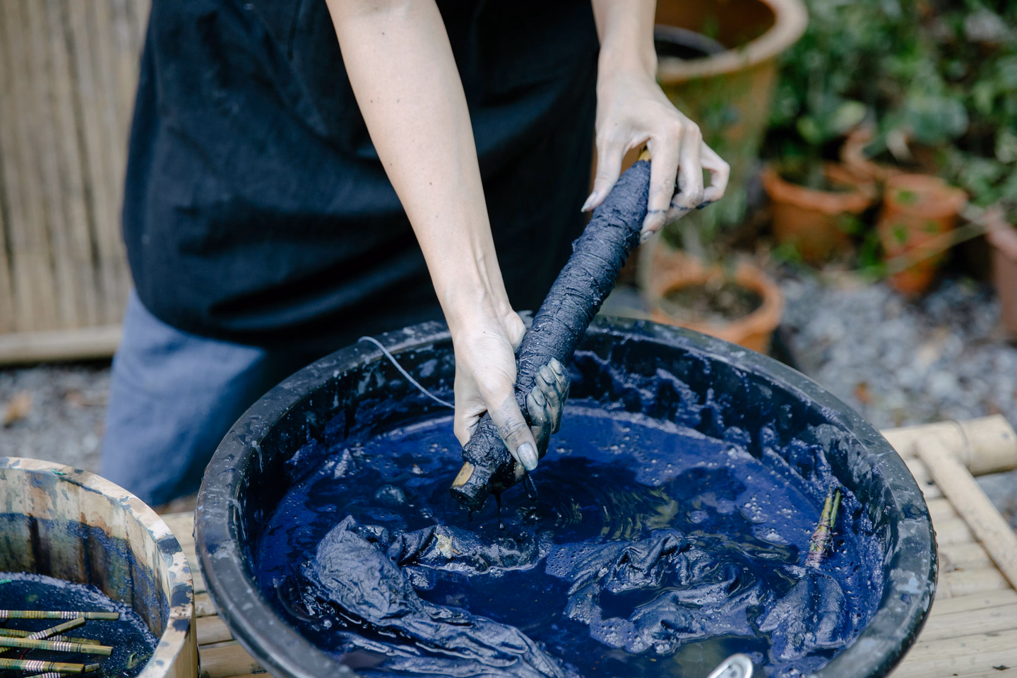 Gloved hands lift a twisted length of fabric out of a black tub of dark blue dye