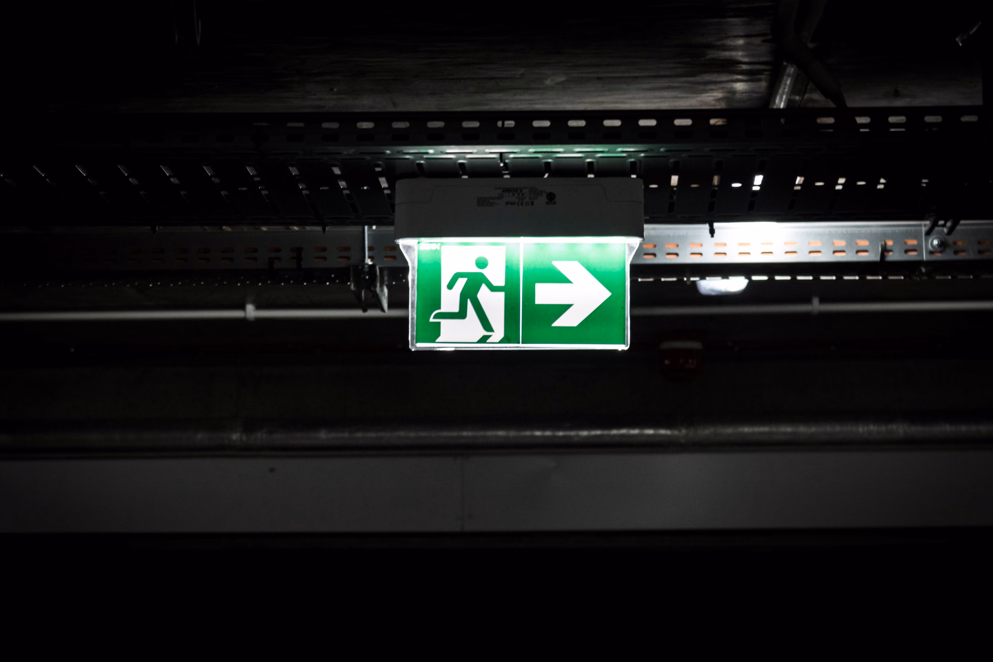 A glowing green emergency exit sign with an arrow, mounted high in a dark industrial space