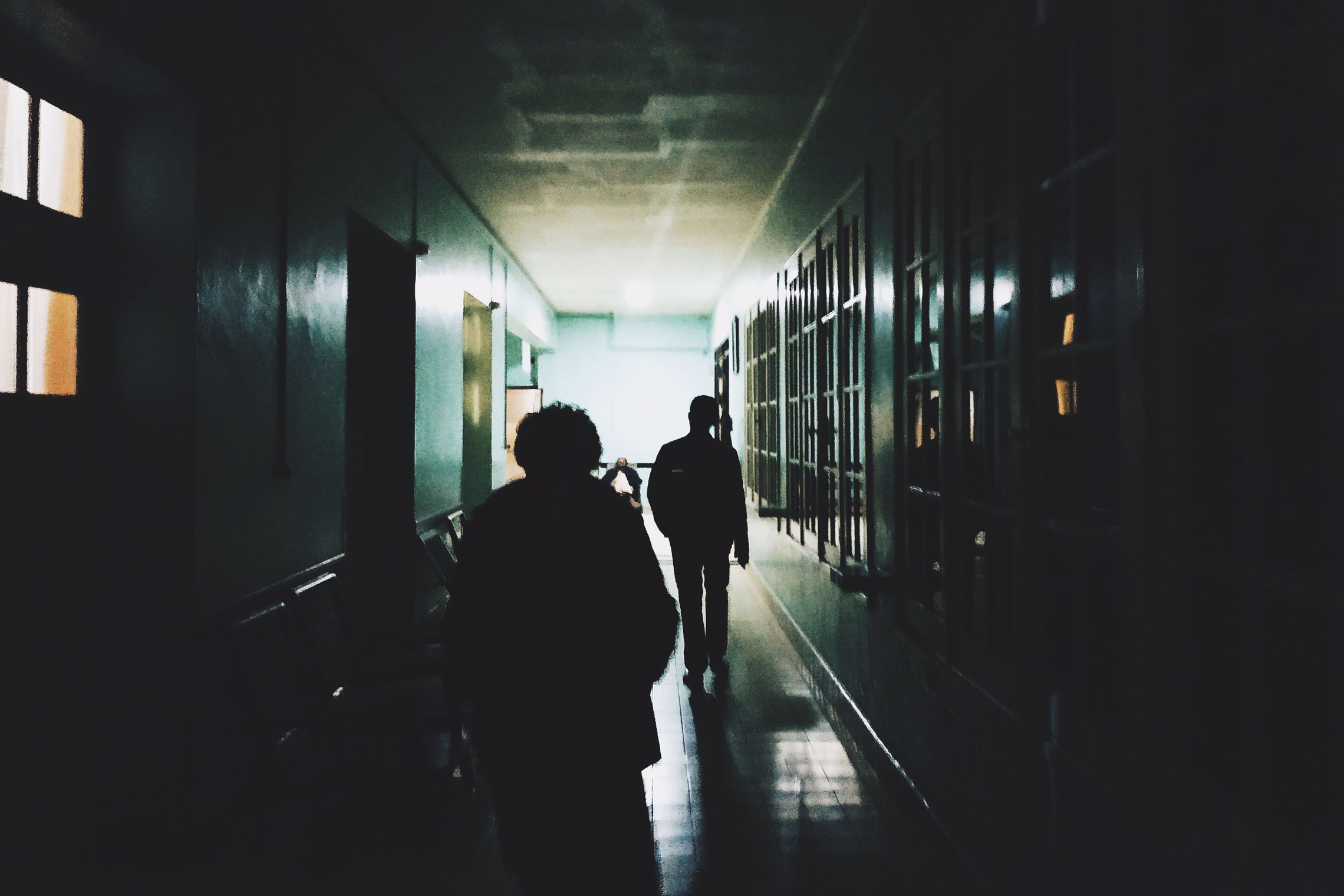 Silhouettes of people walking single file down a long, dim corridor toward a lit doorway