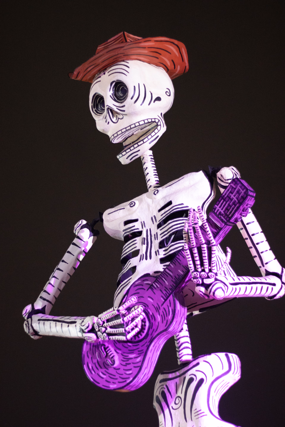 A skeleton figure in a red hat playing a guitar, picked out of the dark by purple light