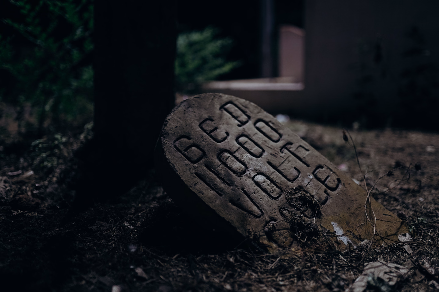 A tilted stone marker with carved lettering catching the light on dark ground, the yard around it in shadow