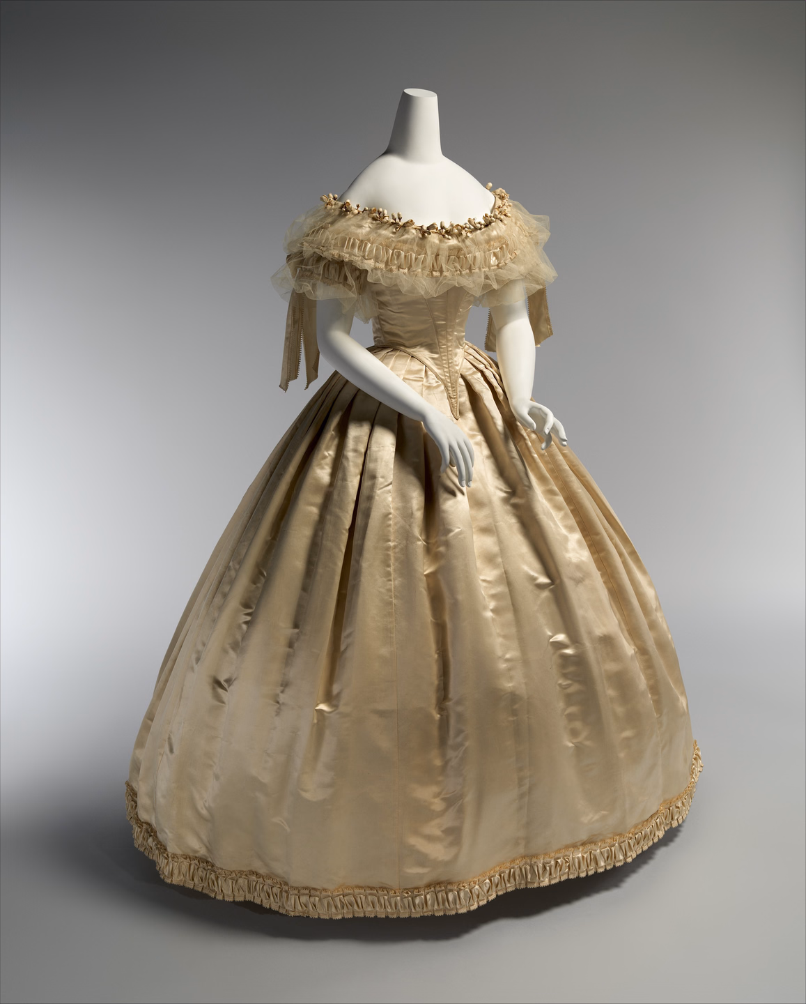 A cream silk wedding dress on a headless mannequin: off-the-shoulder neckline trimmed with sheer tulle and small fabric flowers, a fitted pointed bodice and a very full, pleated bell skirt with a ruched band at the hem