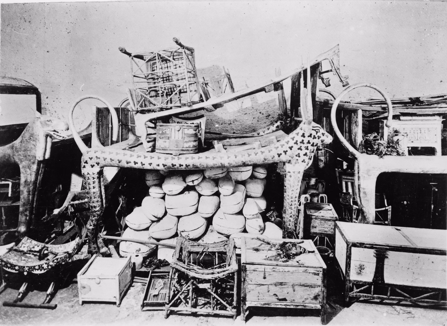 Black and white photograph of a crowded tomb room: a long couch with animal-shaped sides and cow horns, its body painted with a clover-like pattern, stacked under and around with white oval boxes, wooden chests, stools, a folded bed and other furniture against a pale wall