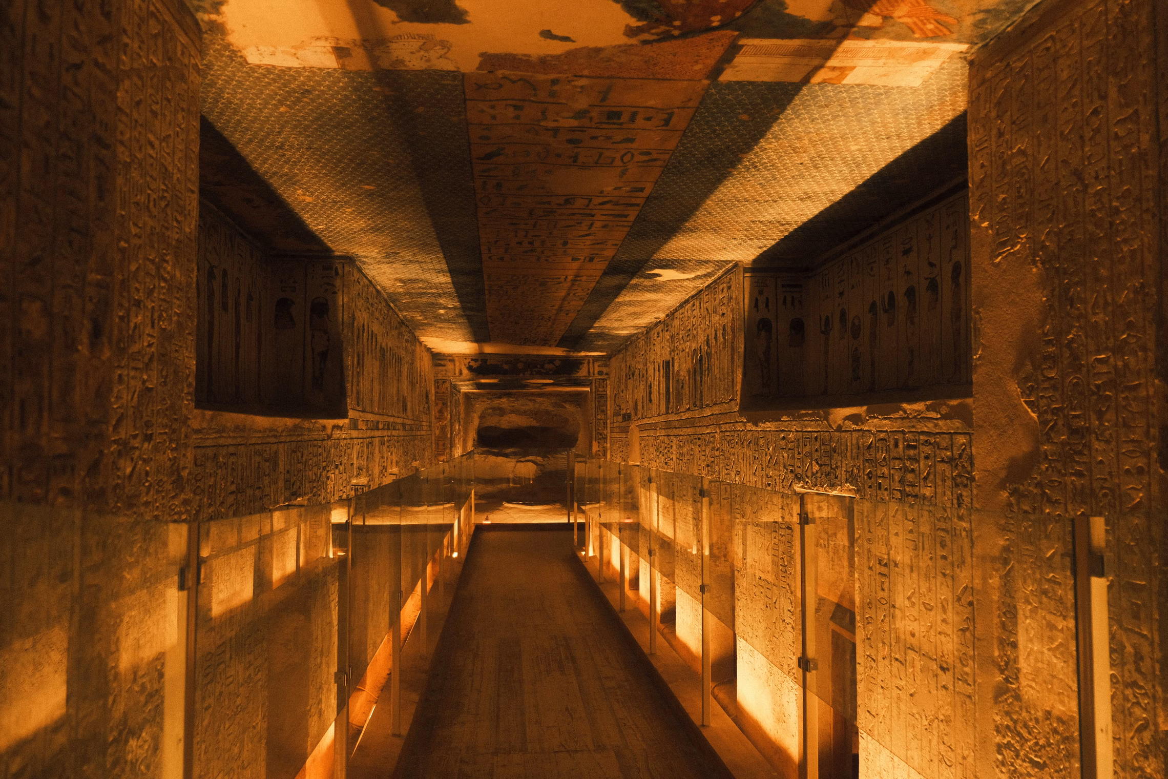 A long, dim tomb corridor with carved and painted walls and ceiling, lit low and orange from the floor, running back to a small doorway at the far end