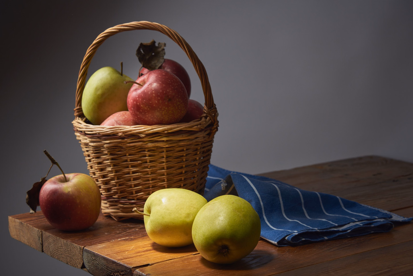 A small wicker basket of red and green apples and a pear on a wooden board, with more apples and two green pears on the board beside a folded blue striped cloth, against a grey wall