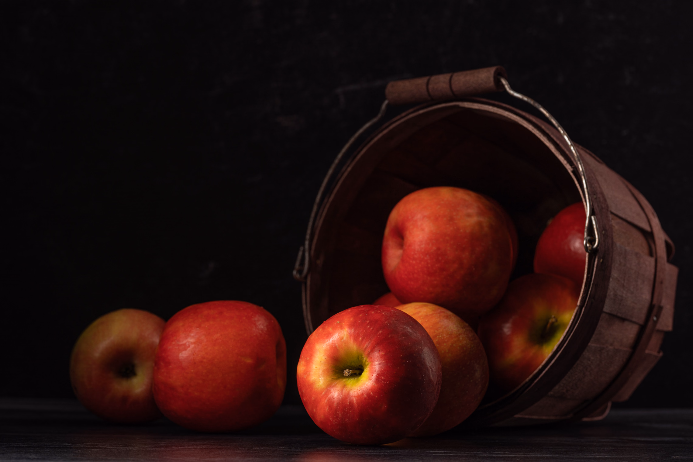 Red apples spilling out of a tipped wooden bushel basket onto a dark table, lit low from one side against a black background